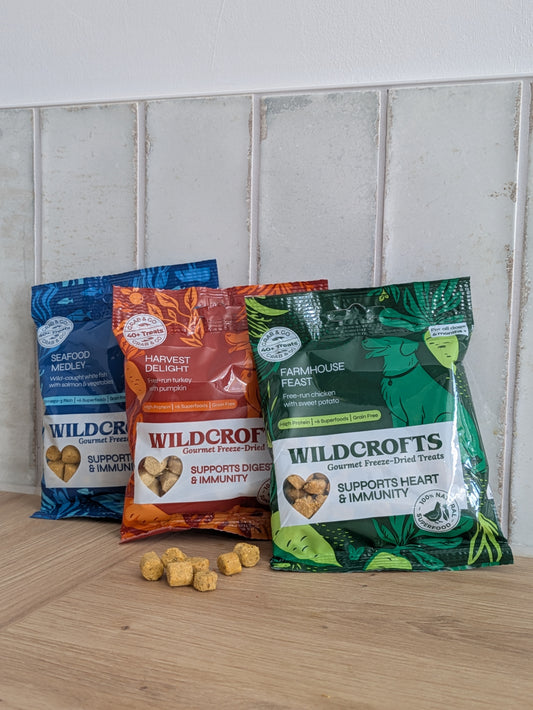 Three bags of Wildcrofts gourmet freeze-dried dog treats arranged on a wooden surface against a white tiled wall. The bags are blue, orange, and green, representing different flavors: Seafood Medley, Harvest delight, and Farmhouse Feast.